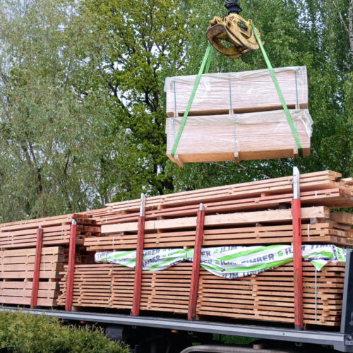 Kran hebt verpackte Paletten mit Schnittholz Schwartenbrettern auf einen beladenen Holztransporter unter grünen Bäumen.
