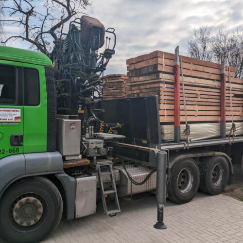 Grüner LKW mit Ladekran transportiert gestapelte Schnittholz Schwartenbretter unter bewölktem Himmel.