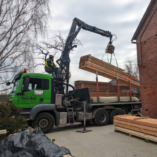 Grüner Voss-Holz LKW entlädt mit Kran Schnittholz Schwartenbretter. Ein Arbeiter steuert den Kran auf dem Hof.