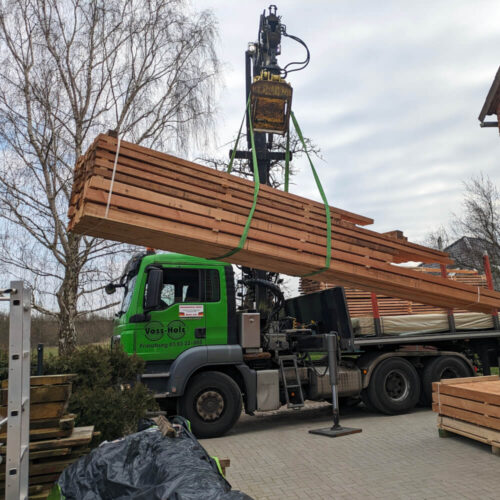 Grüner Voss-Holz LKW hebt großen Stapel Schnittholz Schwartenbretter mit Kran vom Ladebereich.