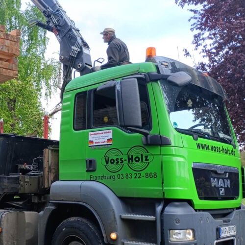 Grüner Voss-Holz LKW mit Kran belädt Schnittholz Schwartenbretter. Ein Arbeiter steuert den Kran sorgfältig.